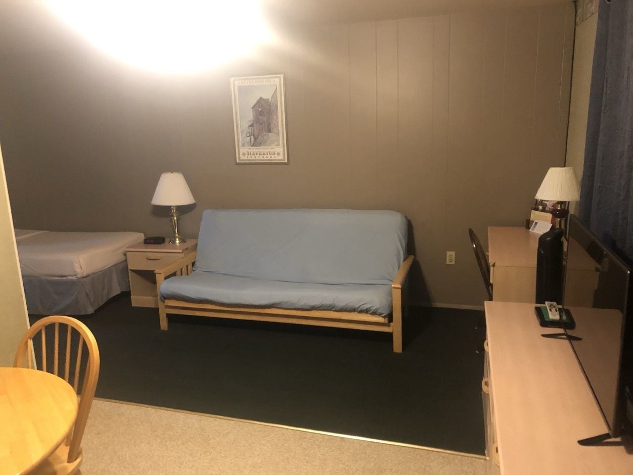 A small, dimly lit room with a single bed, a desk, a chair, and a table.