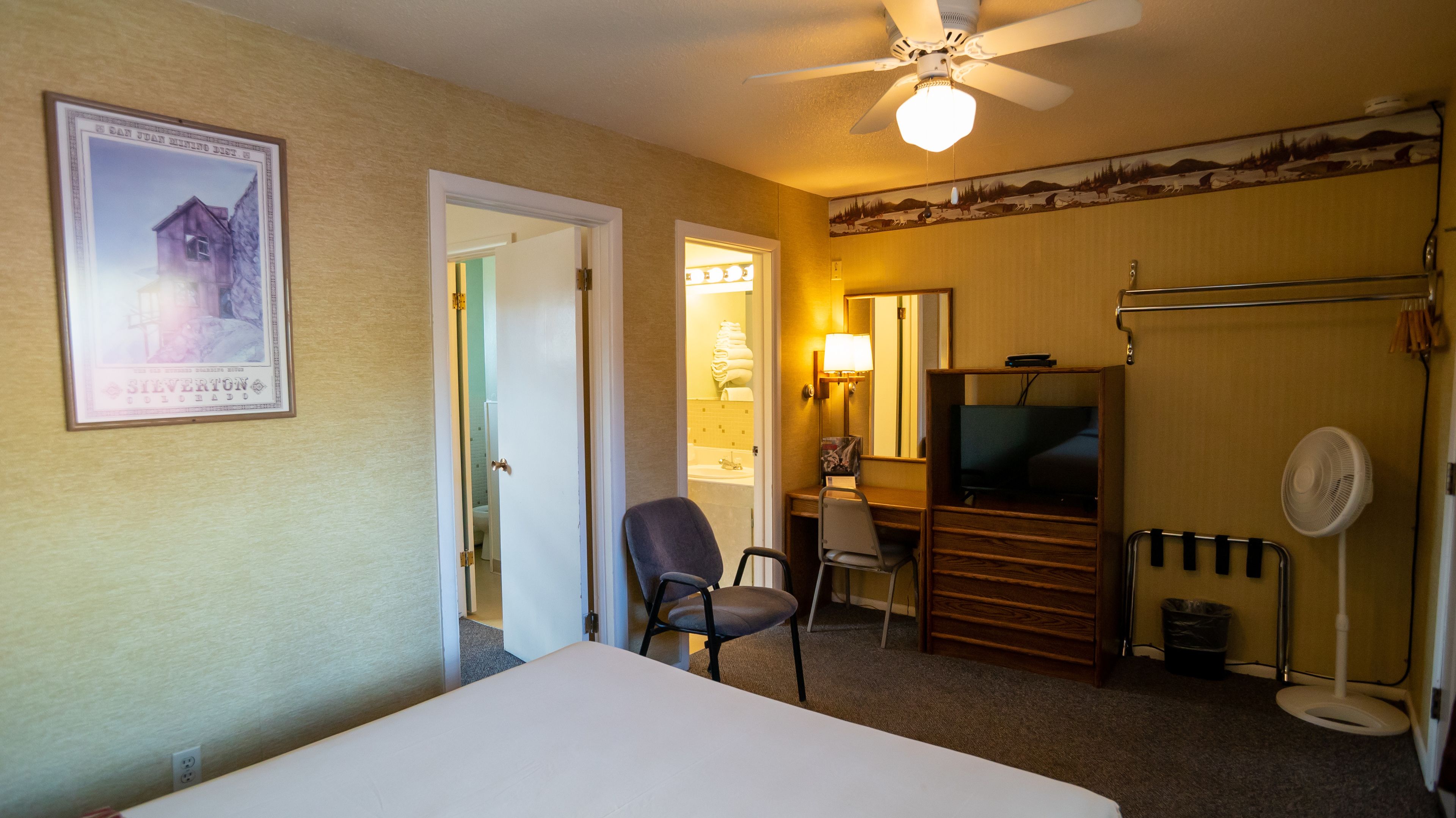 Habitación de hotel con cama, escritorio, silla, televisor, ventilador de techo y baño visible a través de una puerta abierta.