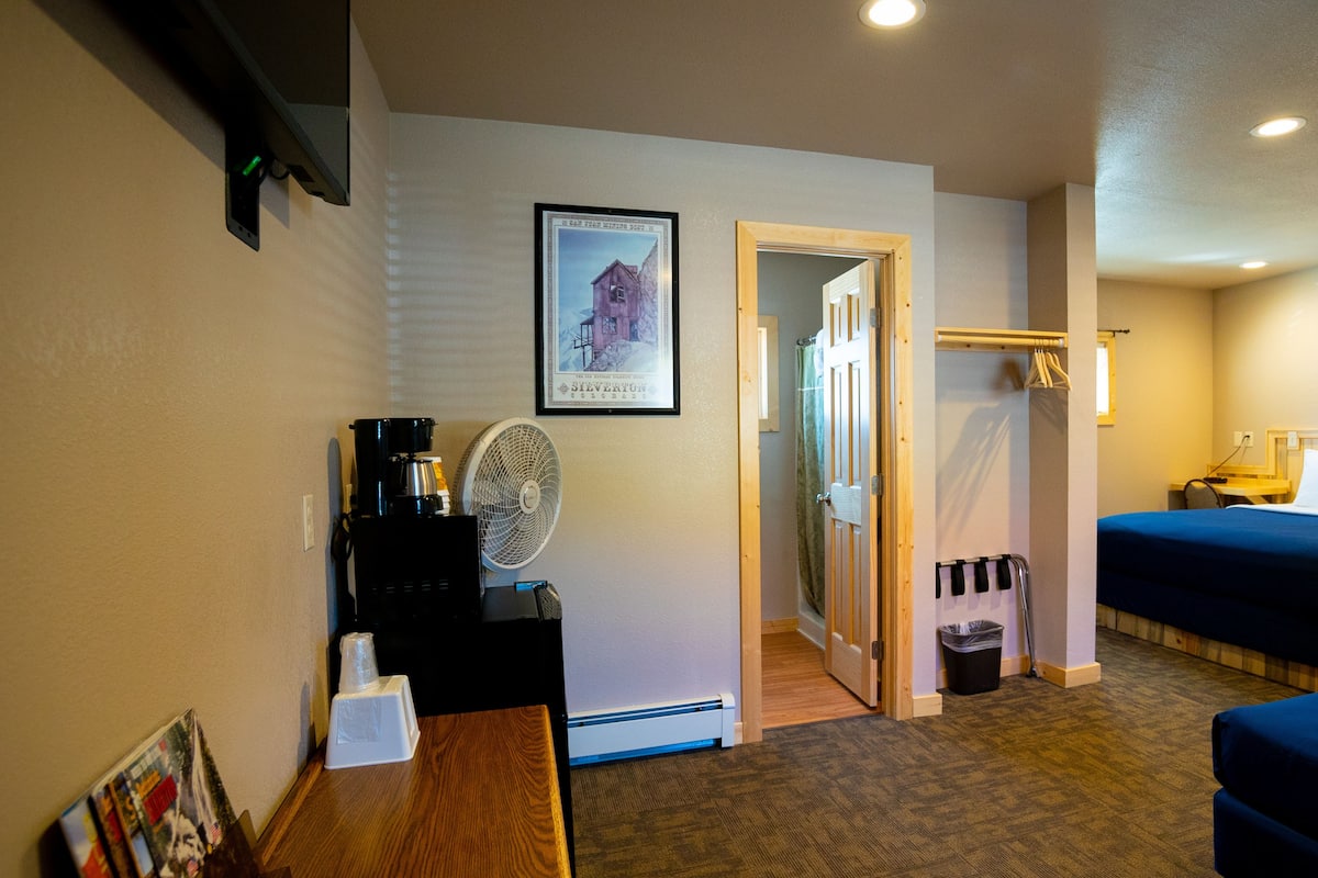 A hotel room with a bed, a desk with a coffee maker, a fan, and a framed picture on the wall.