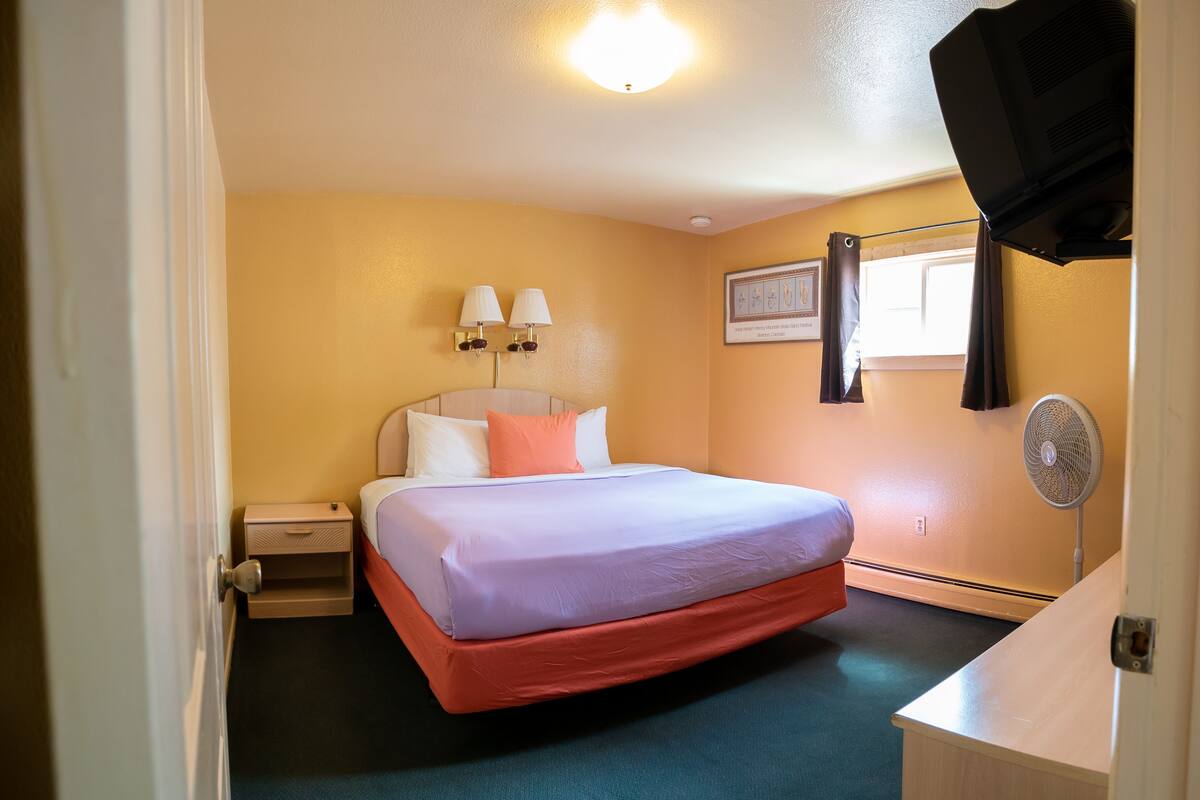 A hotel room with a bed, bedside lamps, a picture on the wall, a fan, and a window with curtains.