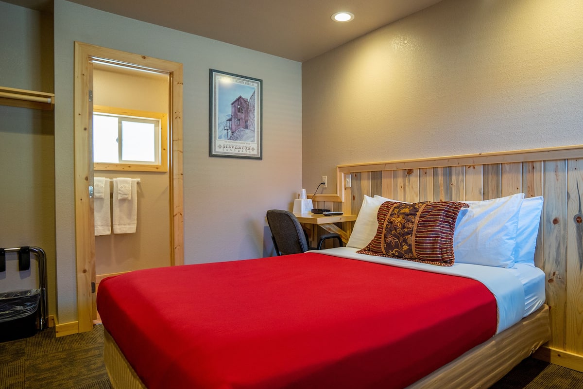 A hotel room with a wooden headboard, a bed with a red bedspread, a desk with a chair, and a framed picture on the wall.