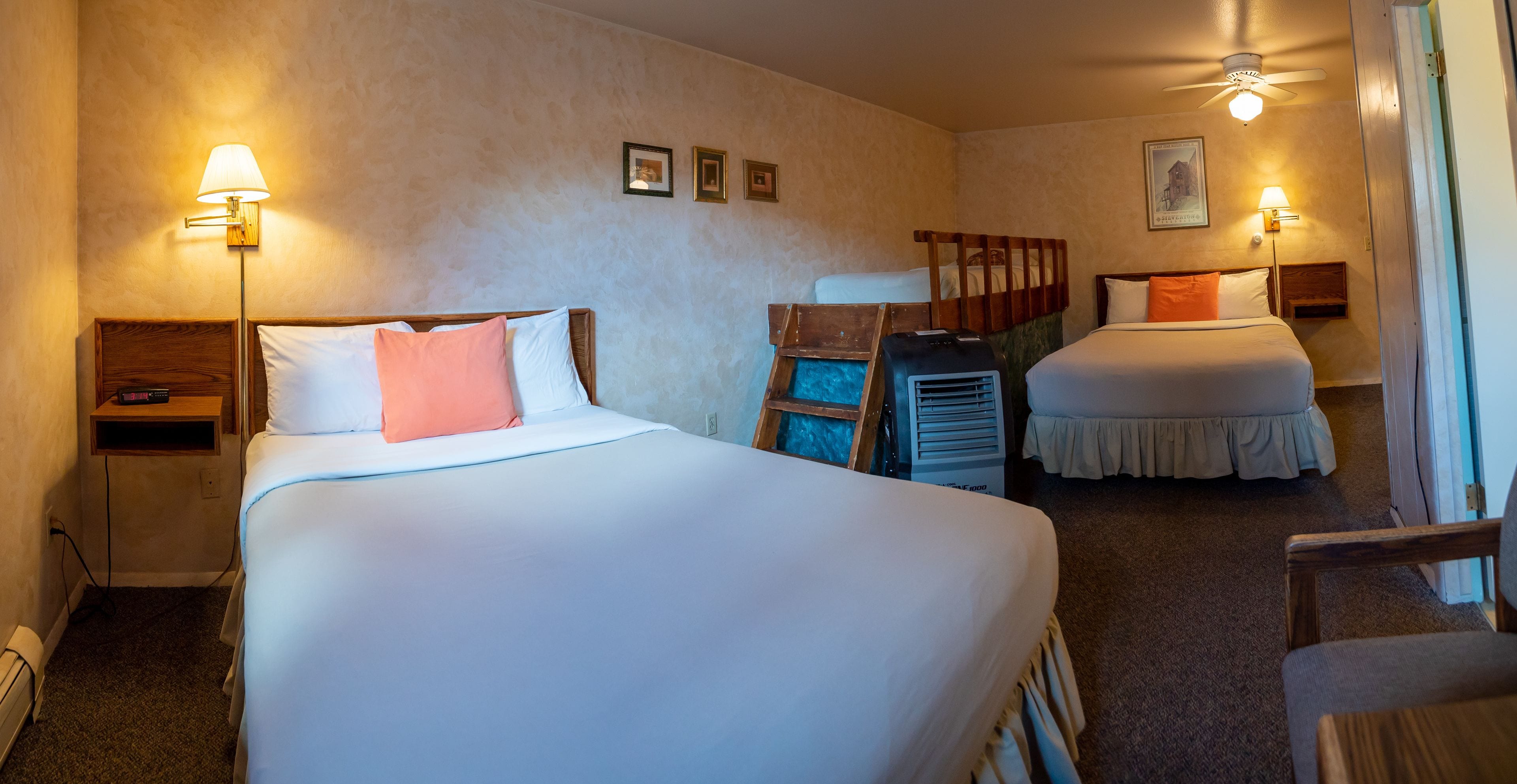 A hotel room with two beds, a desk, a chair, and a ceiling fan.