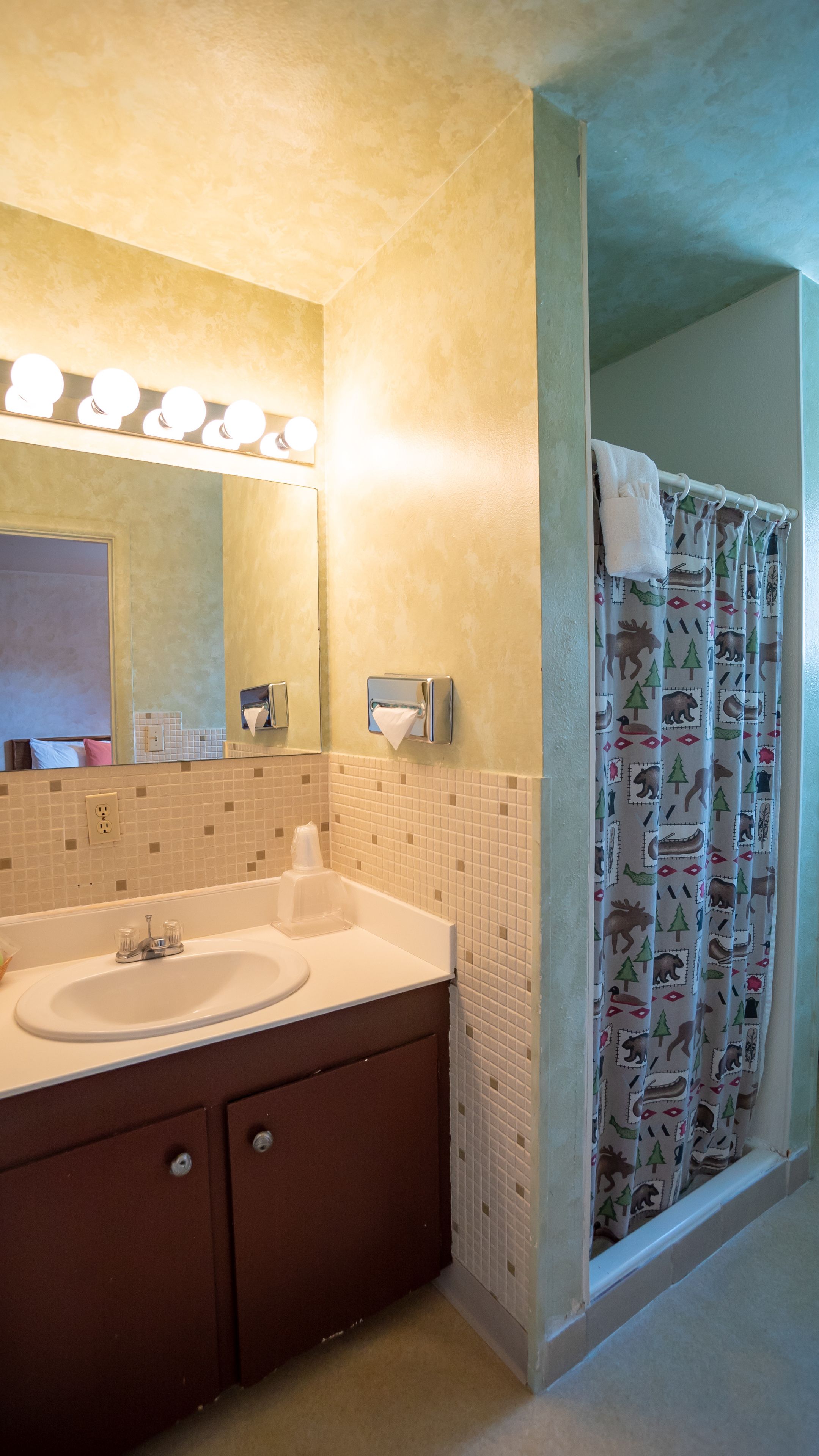 Un baño con lavamanos doble, un espejo con iluminación, un perchero y una cortina de ducha con diseño de bosque.