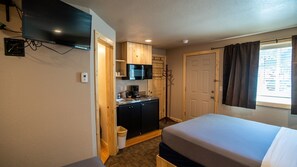 A compact hotel room with a bed, a kitchenette, a TV, and a window with curtains.