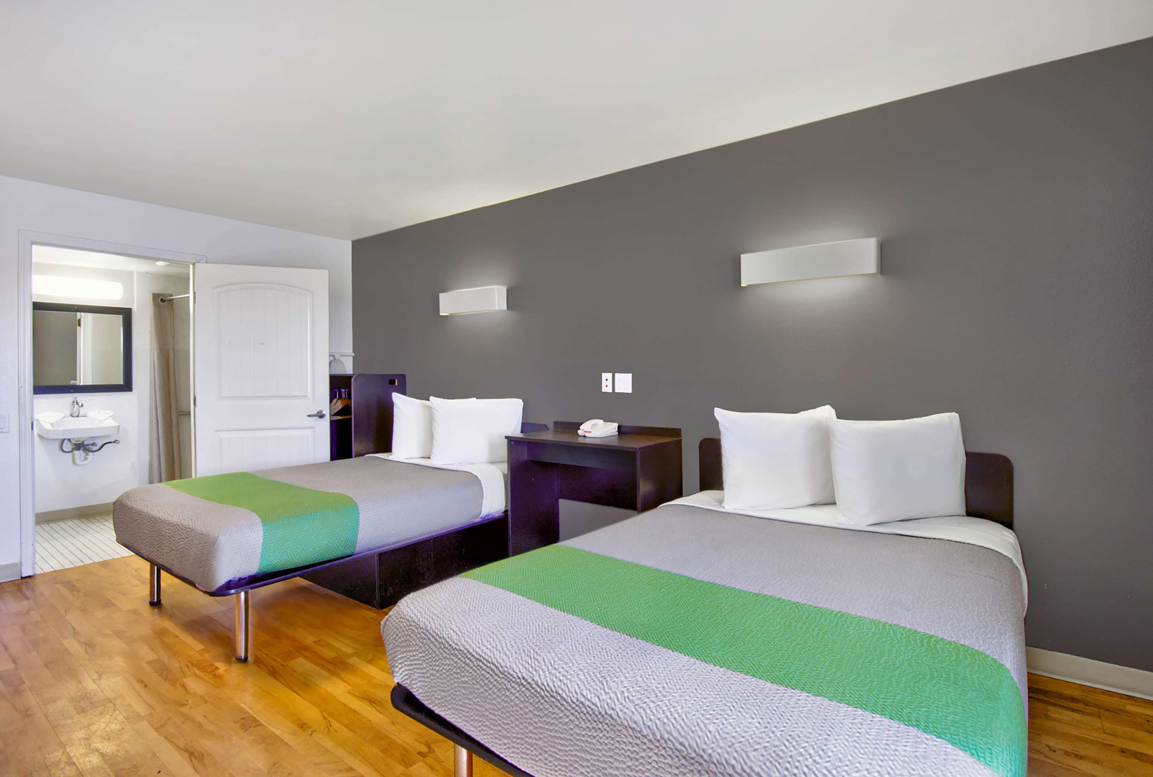 A hotel room with two beds, a bedside table, a nightstand, a mirror, and a bathroom with a sink and mirror.