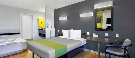 A modern hotel room with a bed, a desk, a chair, a mirror, and a bathroom area.