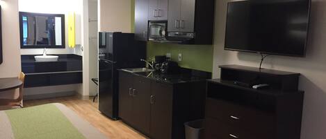 Una cocina moderna con armarios oscuros, un refrigerador negro y un microondas. Hay un pequeño comedor con una silla y una mesa. Un televisor está montado en la pared, y se puede ver un bote de basura.