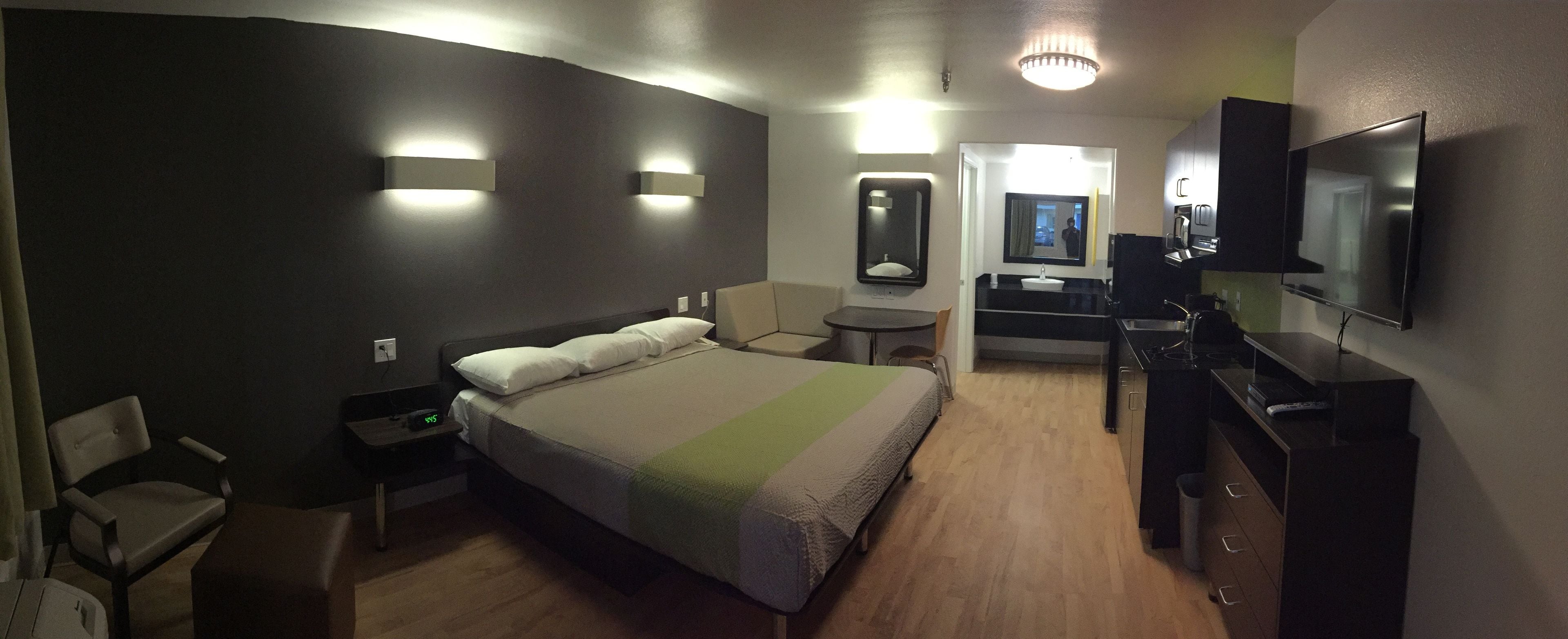 Habitación de hotel con cama, mesita de noche, silla, escritorio, espejo, lavamanos y televisión.