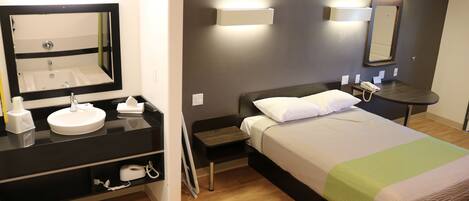 Habitación de hotel con cama, mesita de noche con lámpara, tocador y baño con lavamanos y espejo.