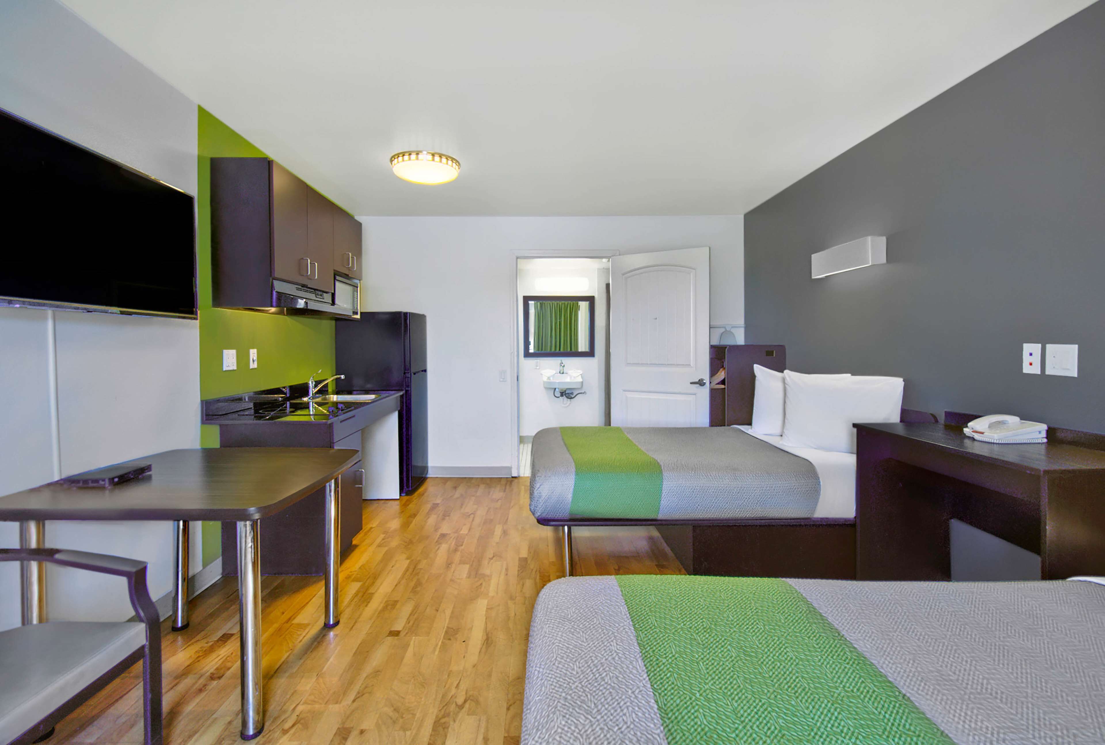Una habitación de hotel compacta con kitchenette, una cama con cubrecama verde y gris, una mesita de noche con un teléfono y un pequeño escritorio con una silla.
