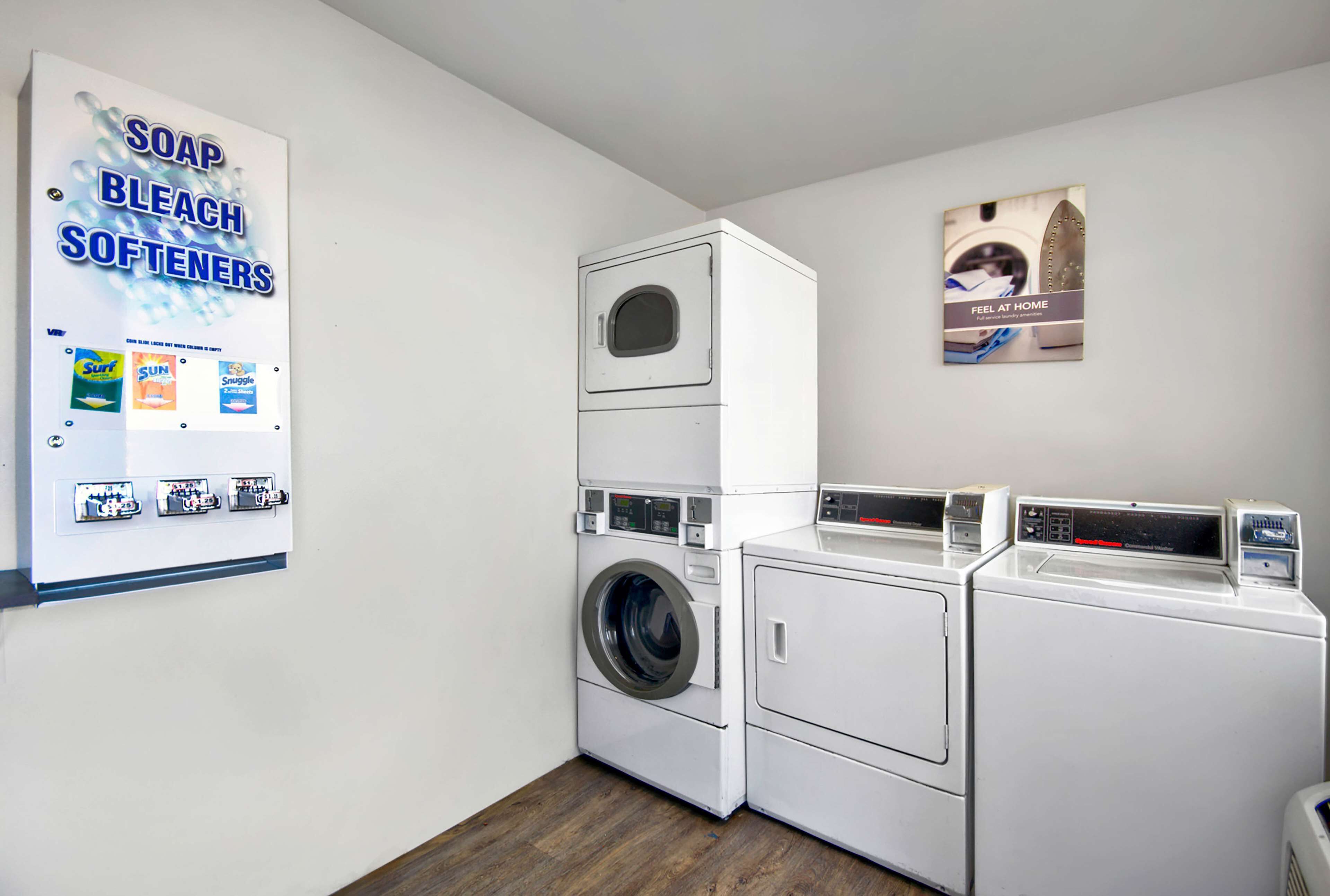 A laundry area with a wall-mounted sign listing soap, bleach, and softeners, and a poster of a person at a home appliance.