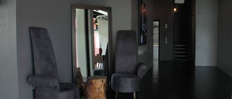A modern lobby with two grey armchairs, a wooden stool, and a large mirror.