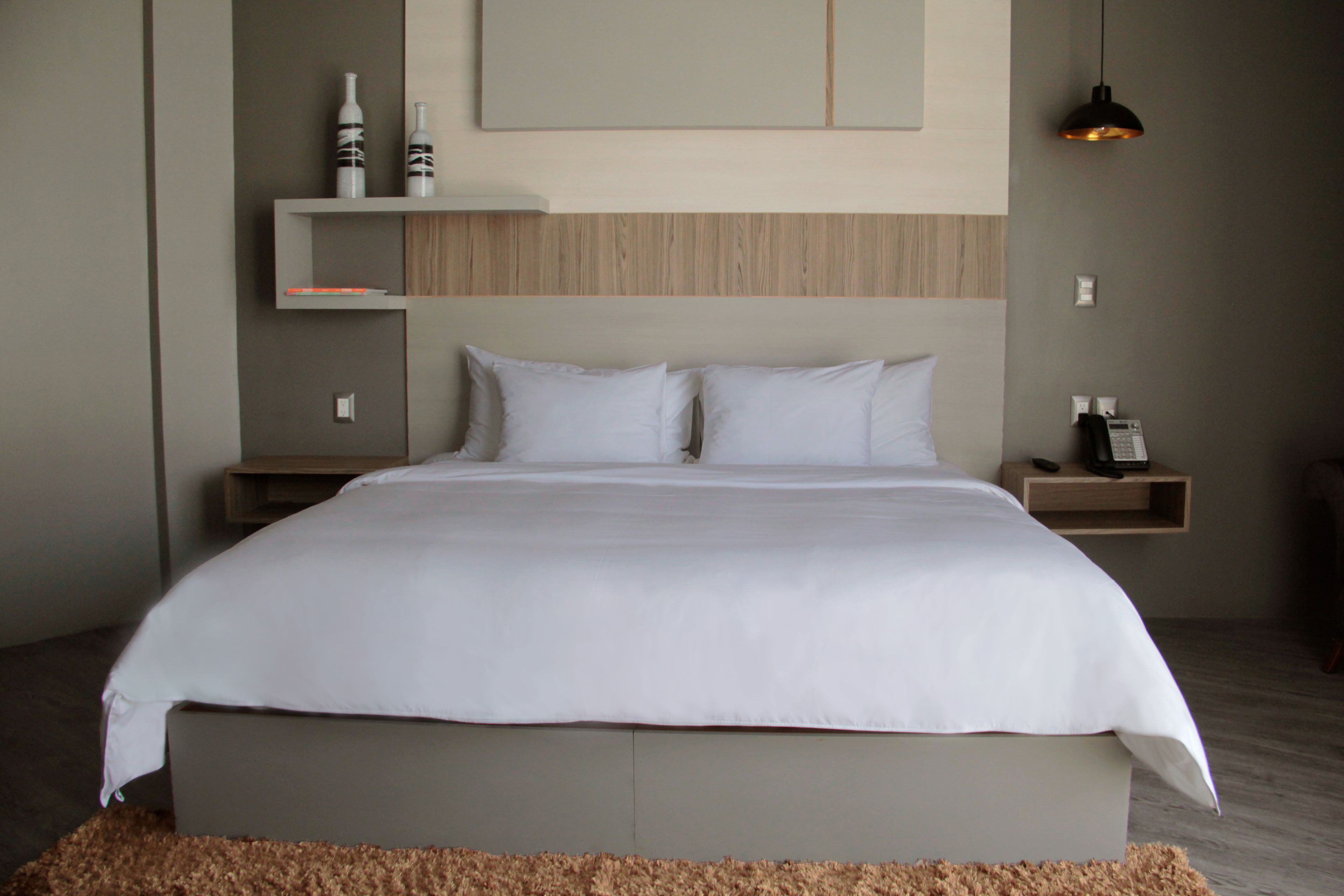 Habitación de hotel moderna con una cama grande, cabecera de madera, mesita de noche con teléfono y un pequeño estante con objetos decorativos.