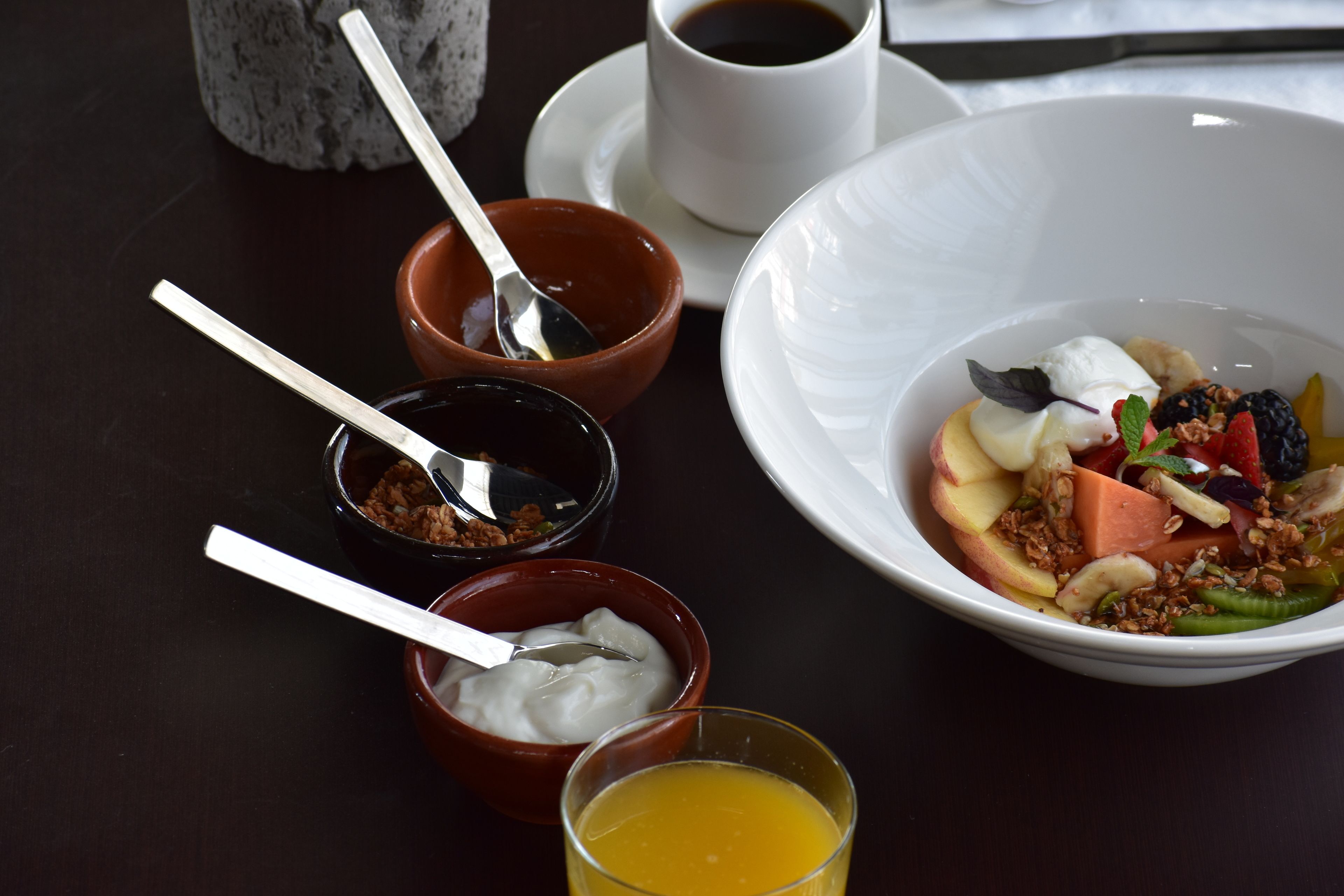 Une table dressée pour le petit-déjeuner, avec un bol de fruits et de yaourt, une tasse de café et un verre de jus d’orange, le tout posé sur une table sombre.