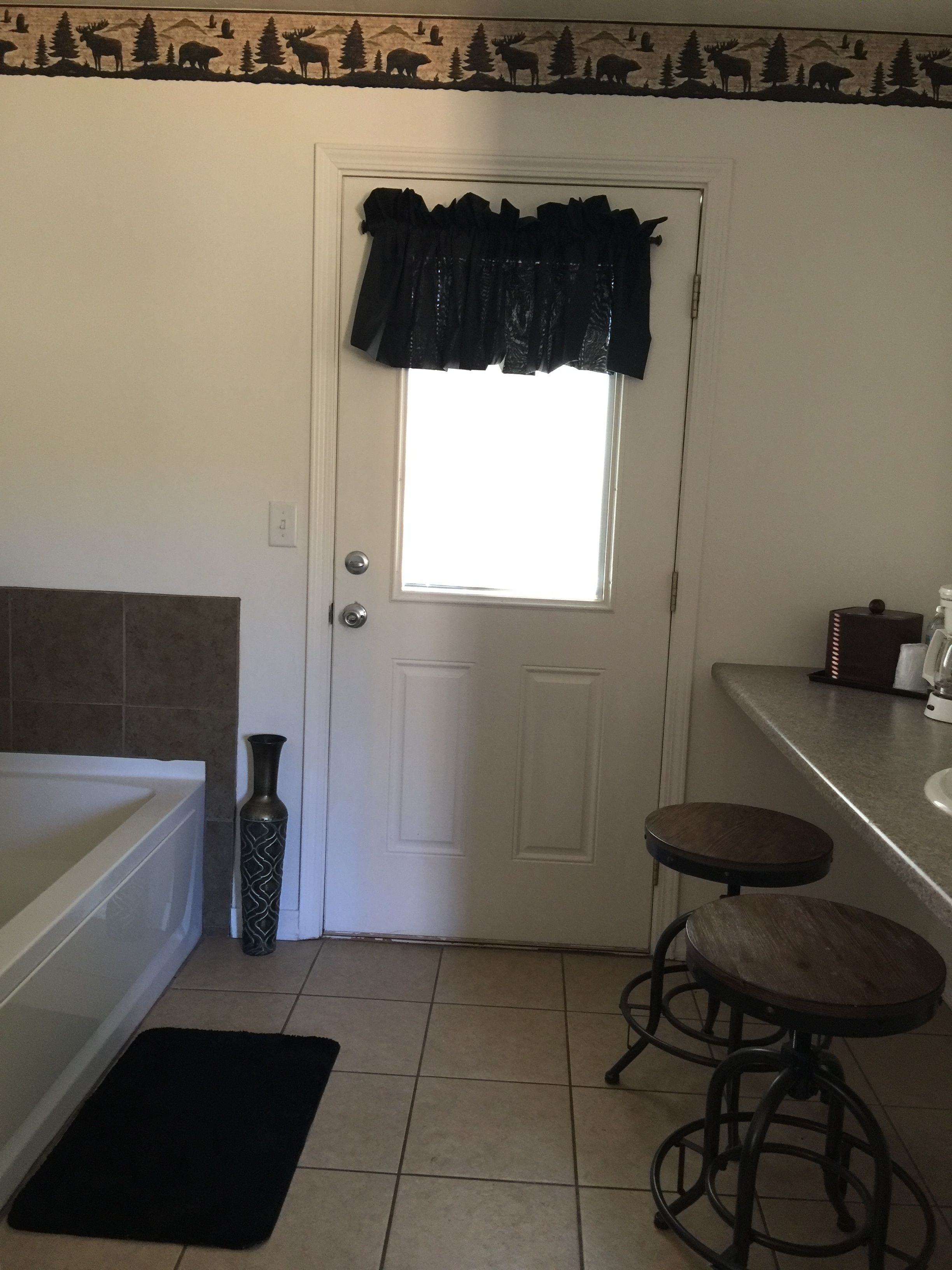 Un baño con una puerta blanca, un ventanal con un valance negro, una bañera, un tapete oscuro y dos taburetes.