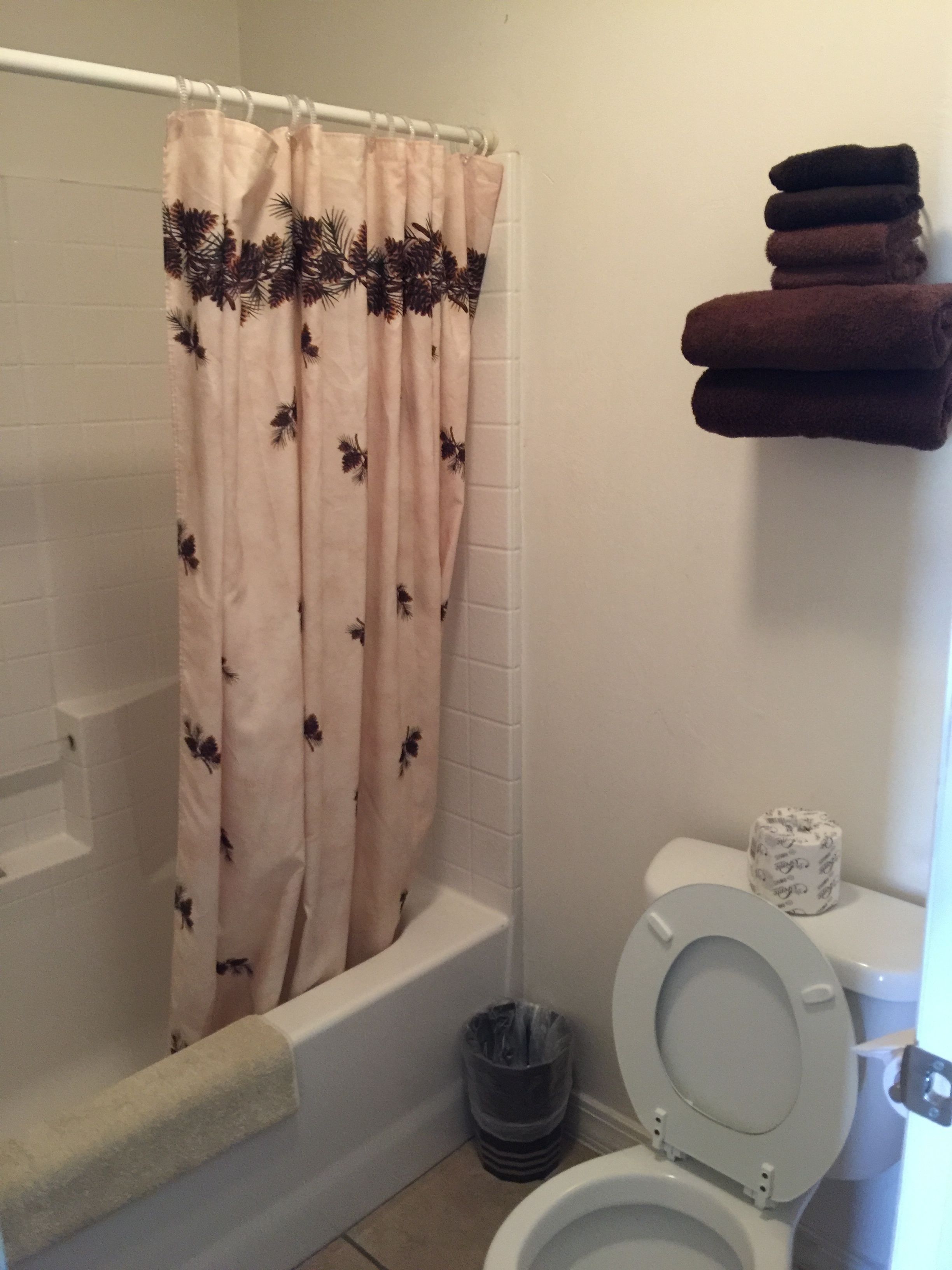 Un baño con cortina de ducha estampada con motivos florales, un inodoro blanco, un cubo de basura y una pila de toallas en un estante.