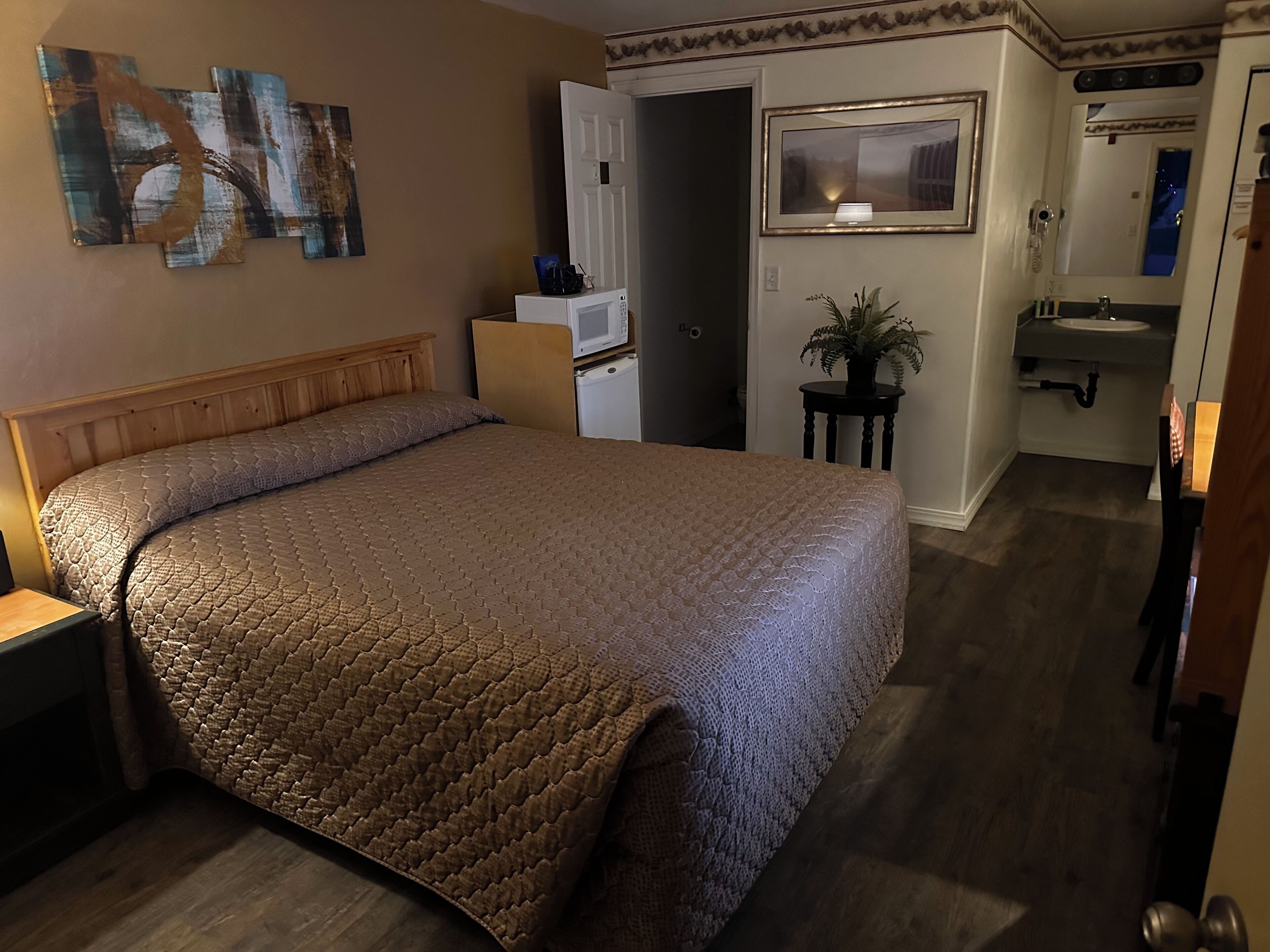 Habitación de hotel con una cama grande, una mesita de noche, un escritorio pequeño, una planta en maceta, un espejo y un baño visible a través de una puerta abierta.