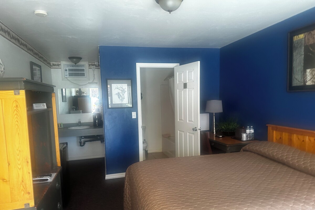 A hotel room with a bed, a desk, a chair, a lamp, a picture frame, and a door leading to a bathroom.