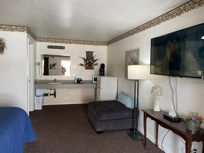 A compact hotel room with a kitchenette, a flat-screen TV, a sofa, and a small table with a lamp and decorative items.