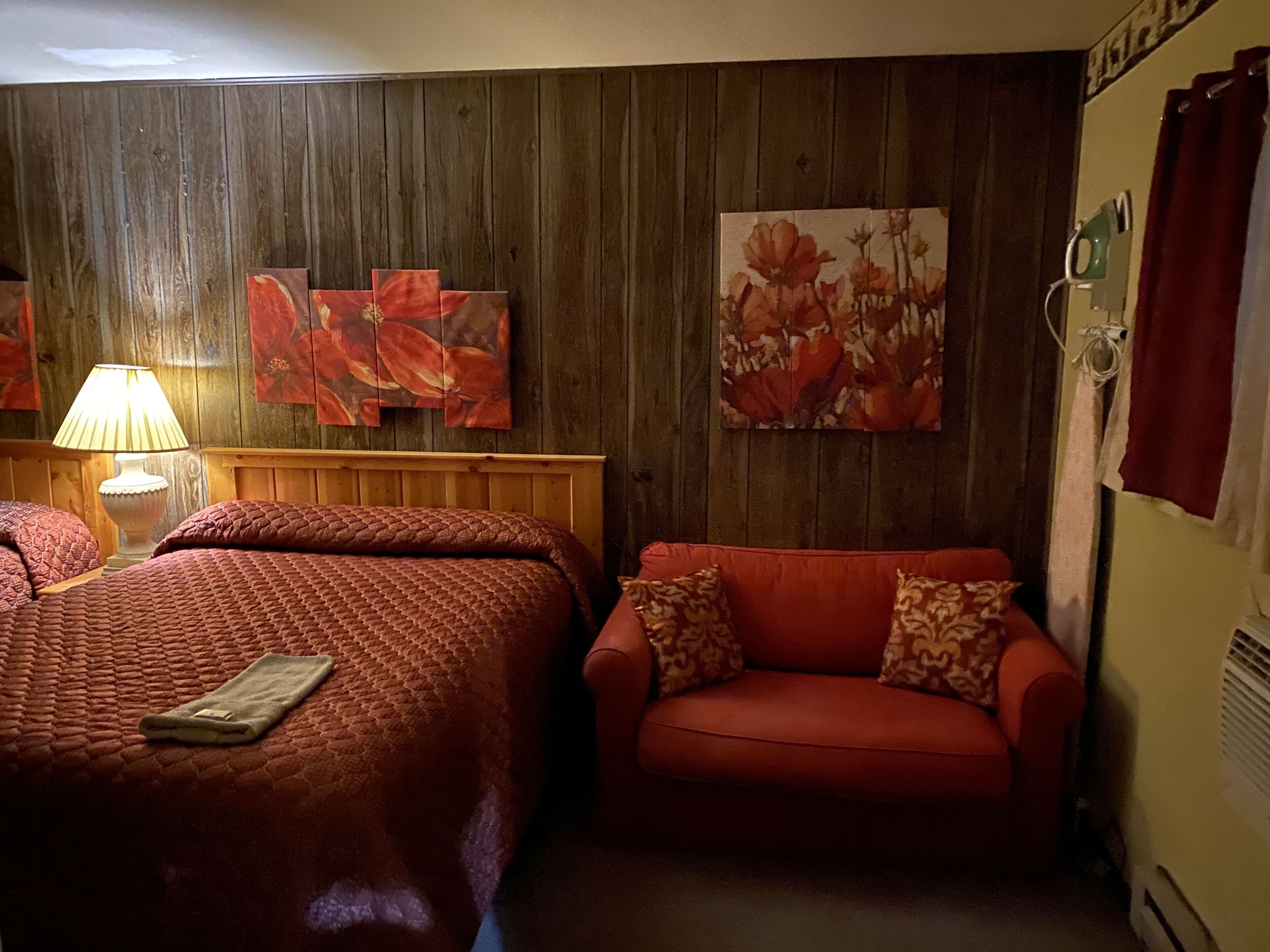 Una habitación con cabecera de madera, un sofá rojo con cojines estampados, una cama con colcha roja y dos cuadros florales en la pared.