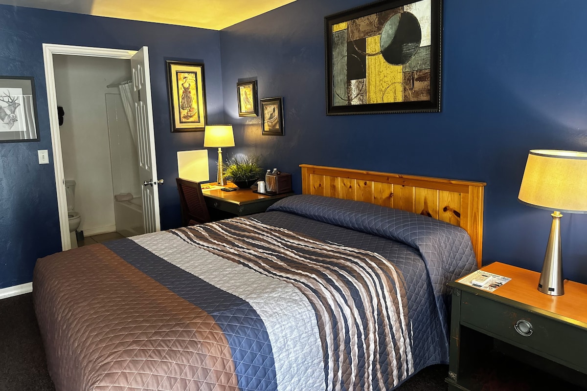 A hotel room with a bed, bedside tables, a desk, a chair, and a bathroom visible through an open door.