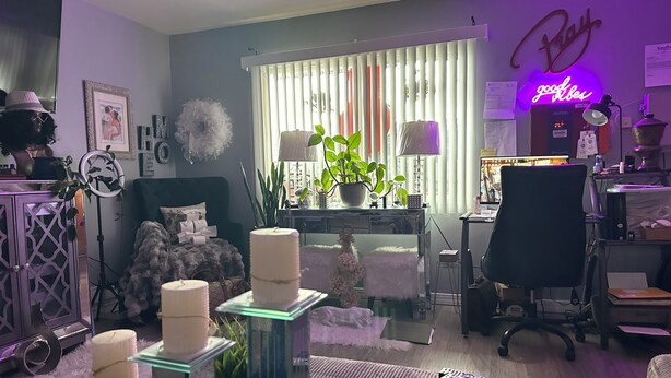 A modern living room with a desk, a comfortable chair, a bed with decorative pillows, a potted plant, and a neon sign on the wall.