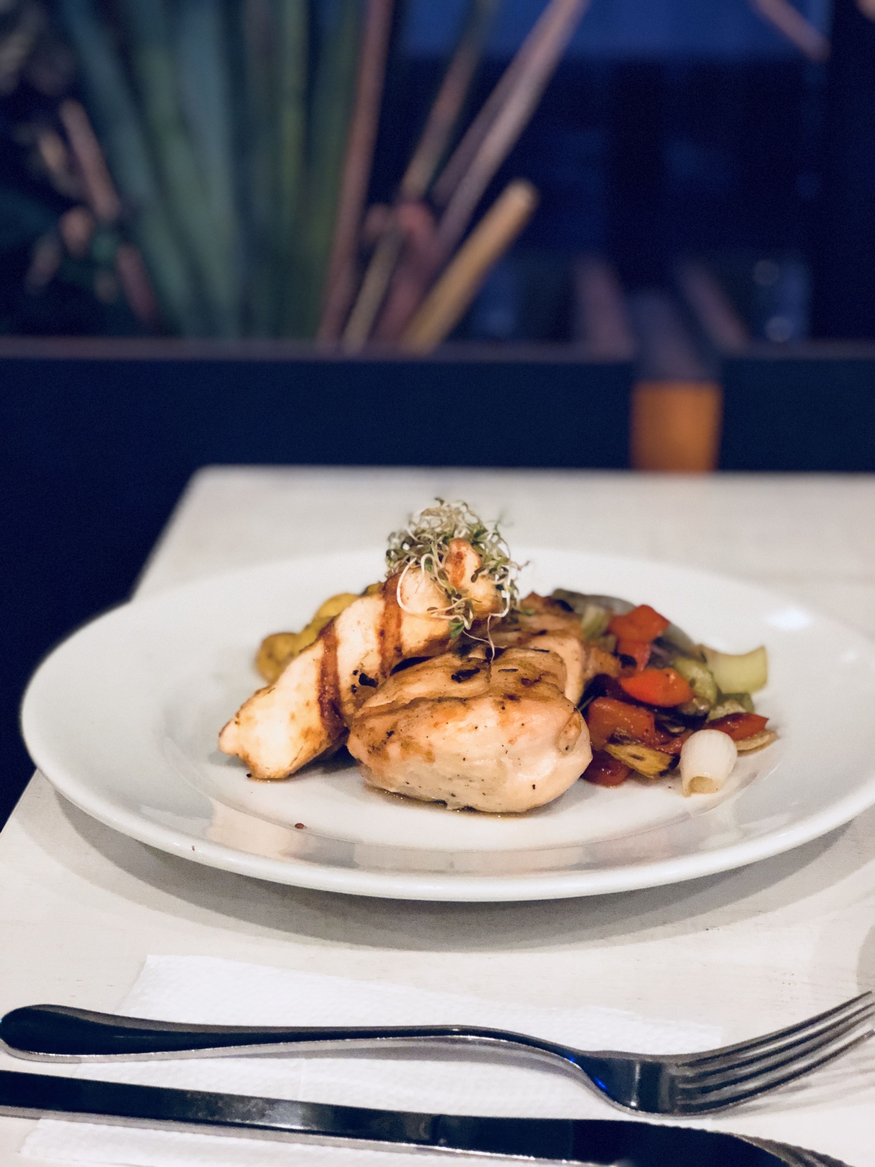 Un plato de pollo a la parrilla con verduras, sobre un plato blanco, colocado en una mesa con un tenedor y un cuchillo a un lado.