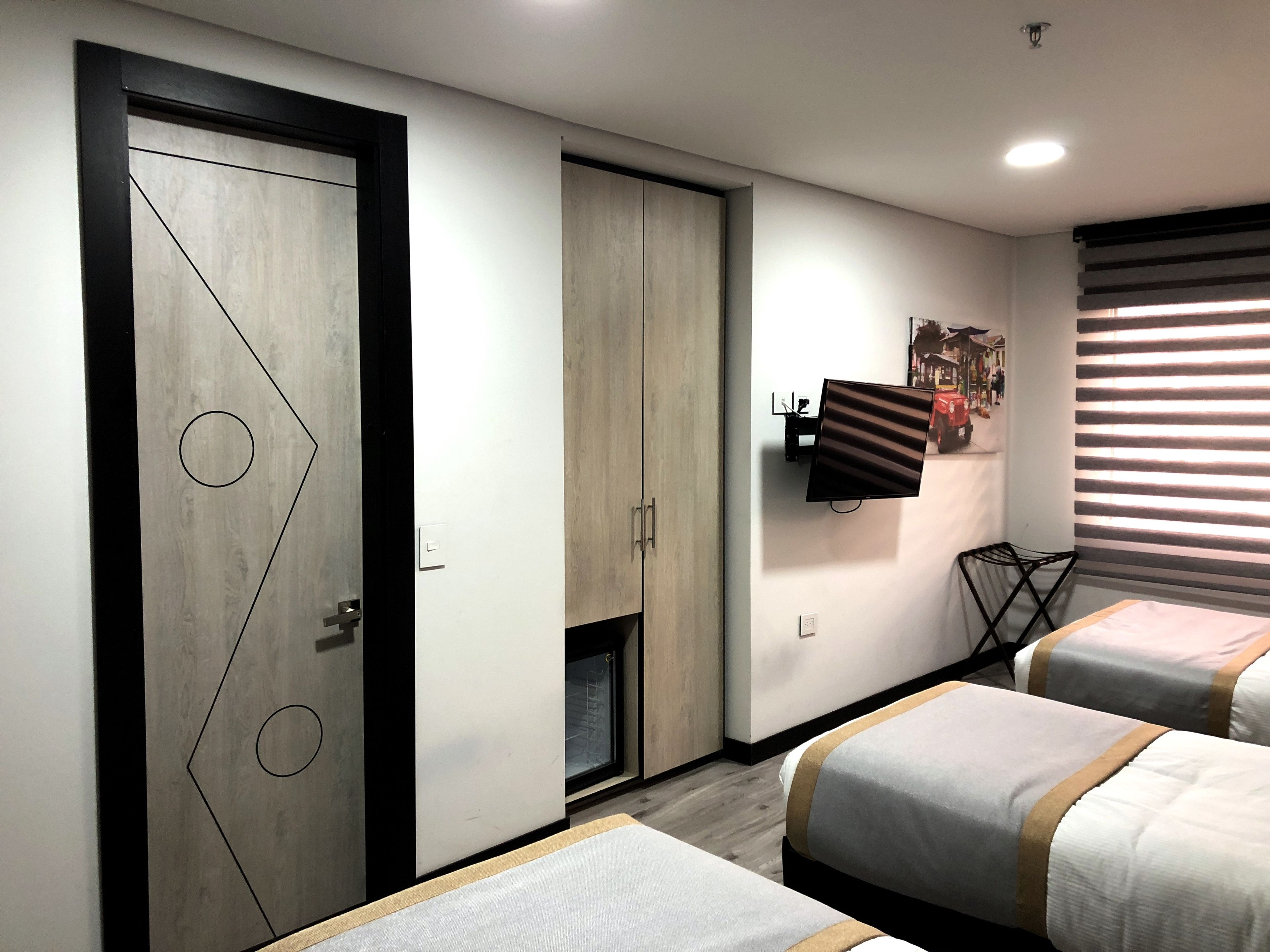A modern bedroom with two beds, a wardrobe, a TV mounted on the wall, and a window with blinds.