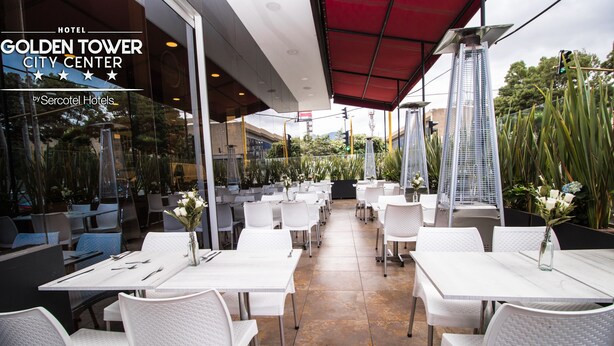 A hotel patio with white tables and chairs, a red awning, and a view of a city center.