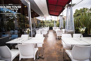 Un patio de hotel con mesas y sillas blancas, un toldo rojo y vistas al centro de la ciudad.