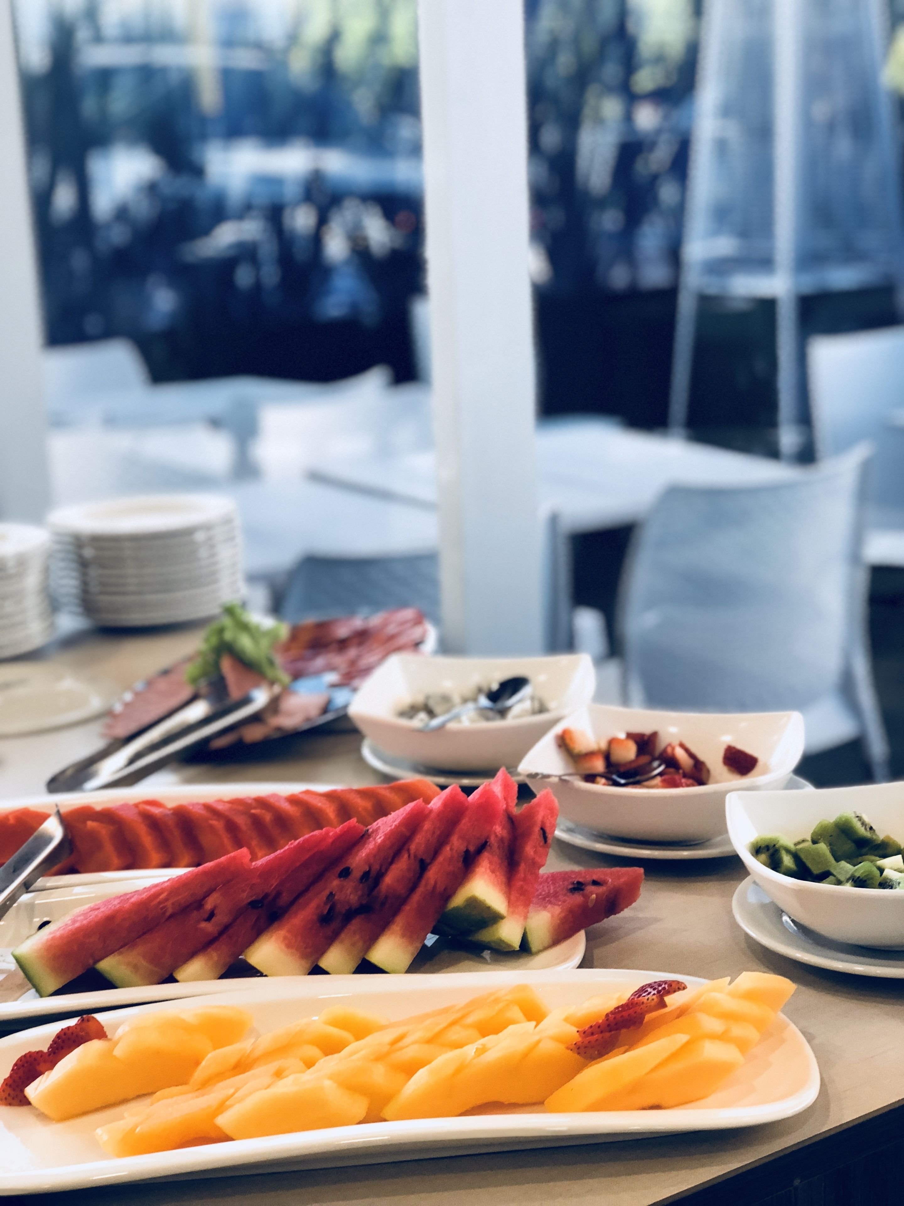 Una mesa con frutas variadas y fuentes de comida, con vista a un edificio y árboles al fondo.