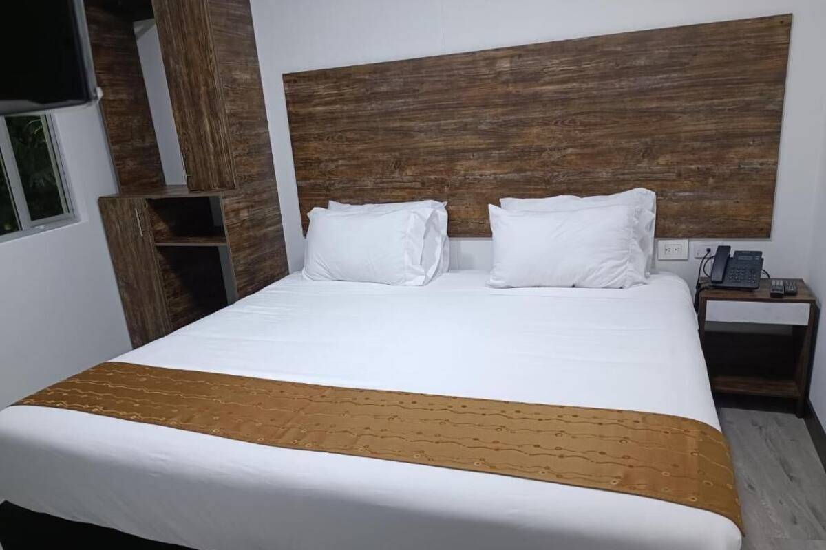 Een net opgemaakt bed met witte lakens en een bruin accent, een houten hoofdbord, een nachtkastje met een telefoon, en een klein rekje.