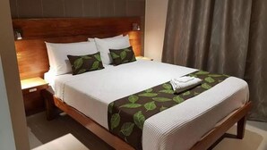 Ein ordentlich bezogenes Bett mit Holz-Kopfschild, weißen Leinen und einer Decke mit grünem Blattmuster.