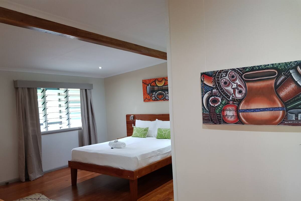 A hotel room with a wooden bed, a window with blinds, a painting on the wall, and a woven rug on the floor.