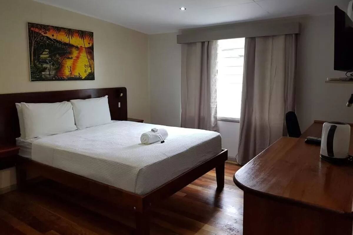 A hotel room with a bed, a desk, a TV, and a painting on the wall.