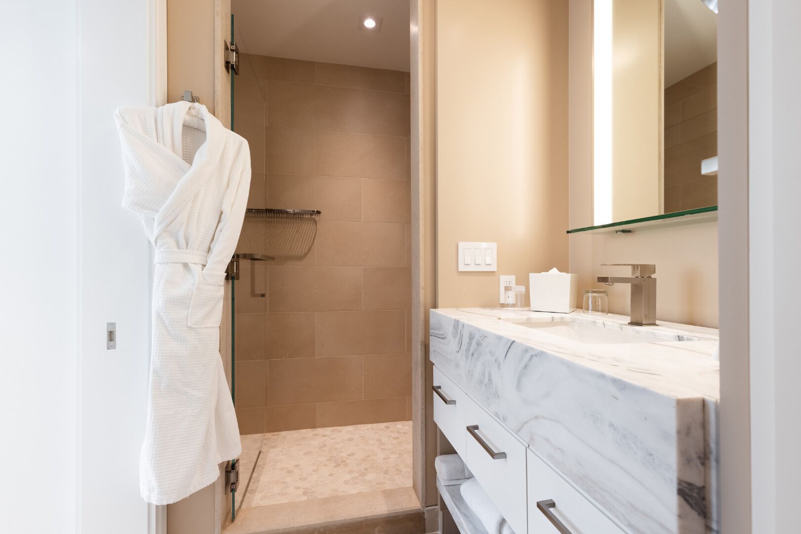 A modern bathroom with a walk-in shower, a marble vanity, and a white robe hanging on a rack.