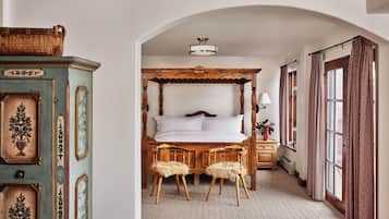 A bedroom with a four-poster bed, two wooden chairs with fur throws, a bedside table with a lamp, and a window with patterned curtains.