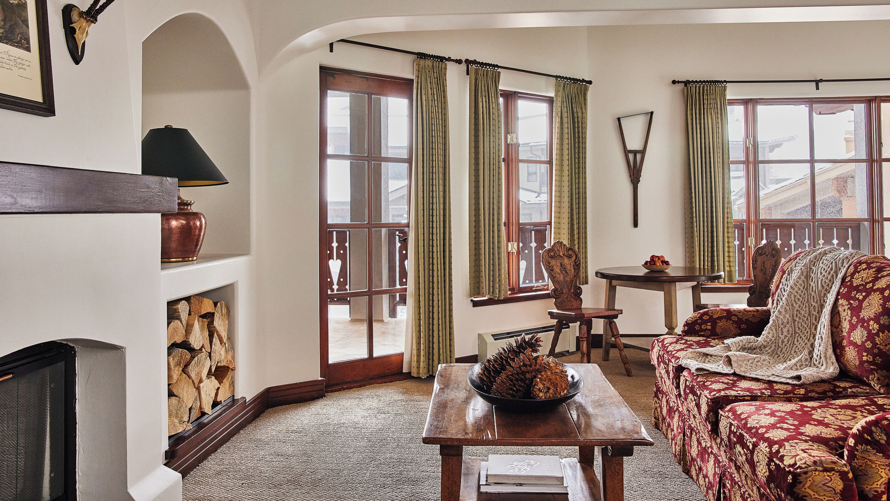 A cozy living room with a fireplace, a wooden table with a bowl of pinecones, a patterned sofa, and a chair.