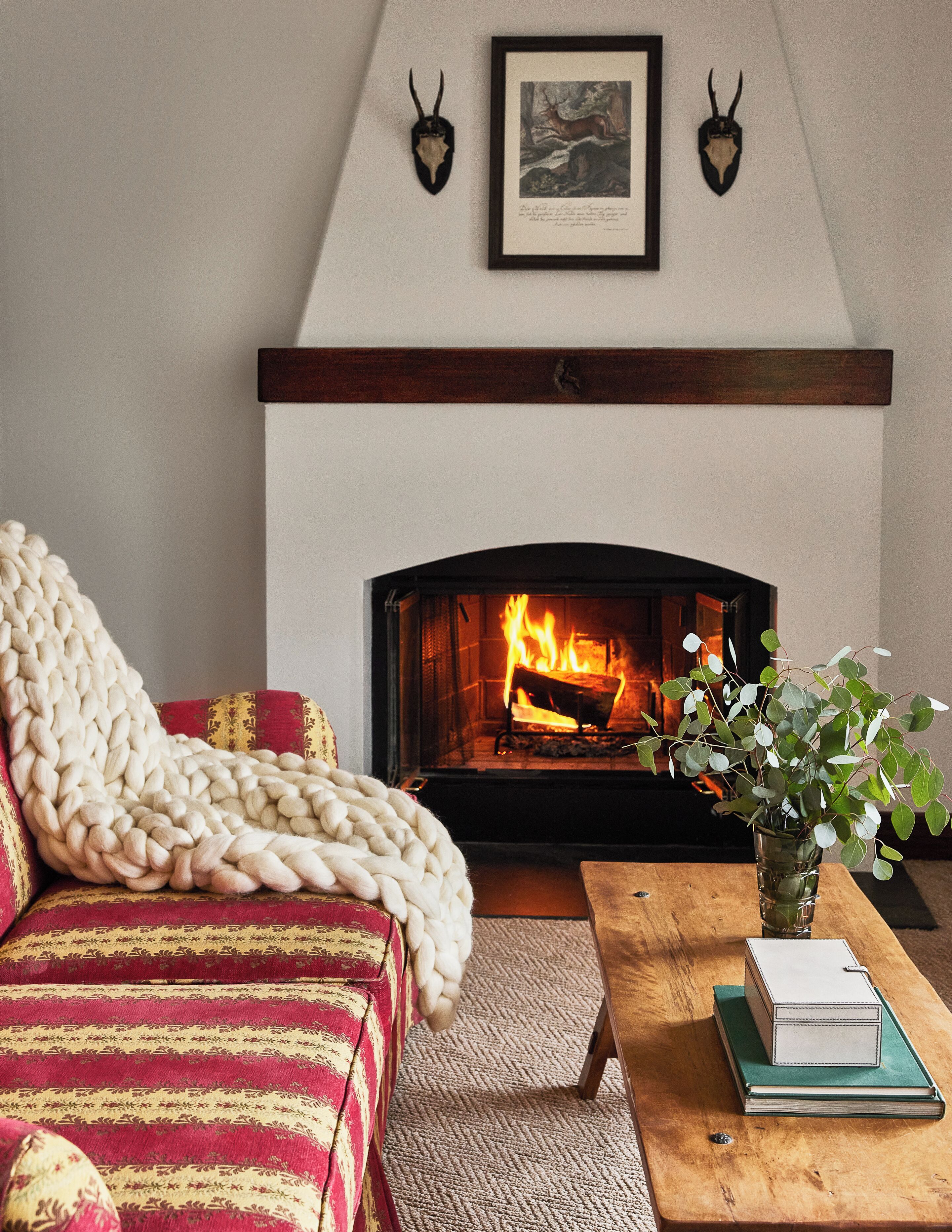 A cozy living room with a fireplace, a red and gold patterned sofa, a chunky knit throw, a wooden coffee table, and a vase with green foliage.