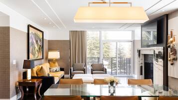 Een moderne hotelkamer met een glazen eettafel, zachte zithoek, een open haard en grote ramen met uitzicht op de omgeving.