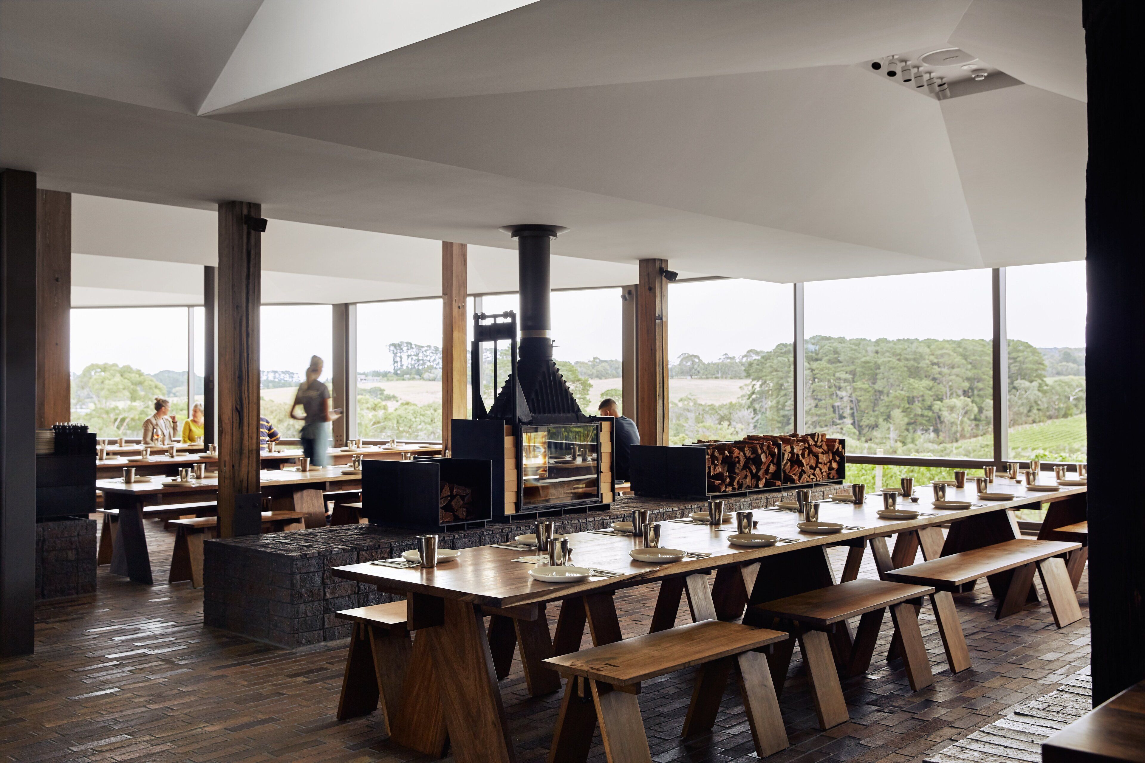 A spacious dining area with high ceilings and large windows, featuring wooden tables and benches, a central fireplace, and a view of the outdoors.