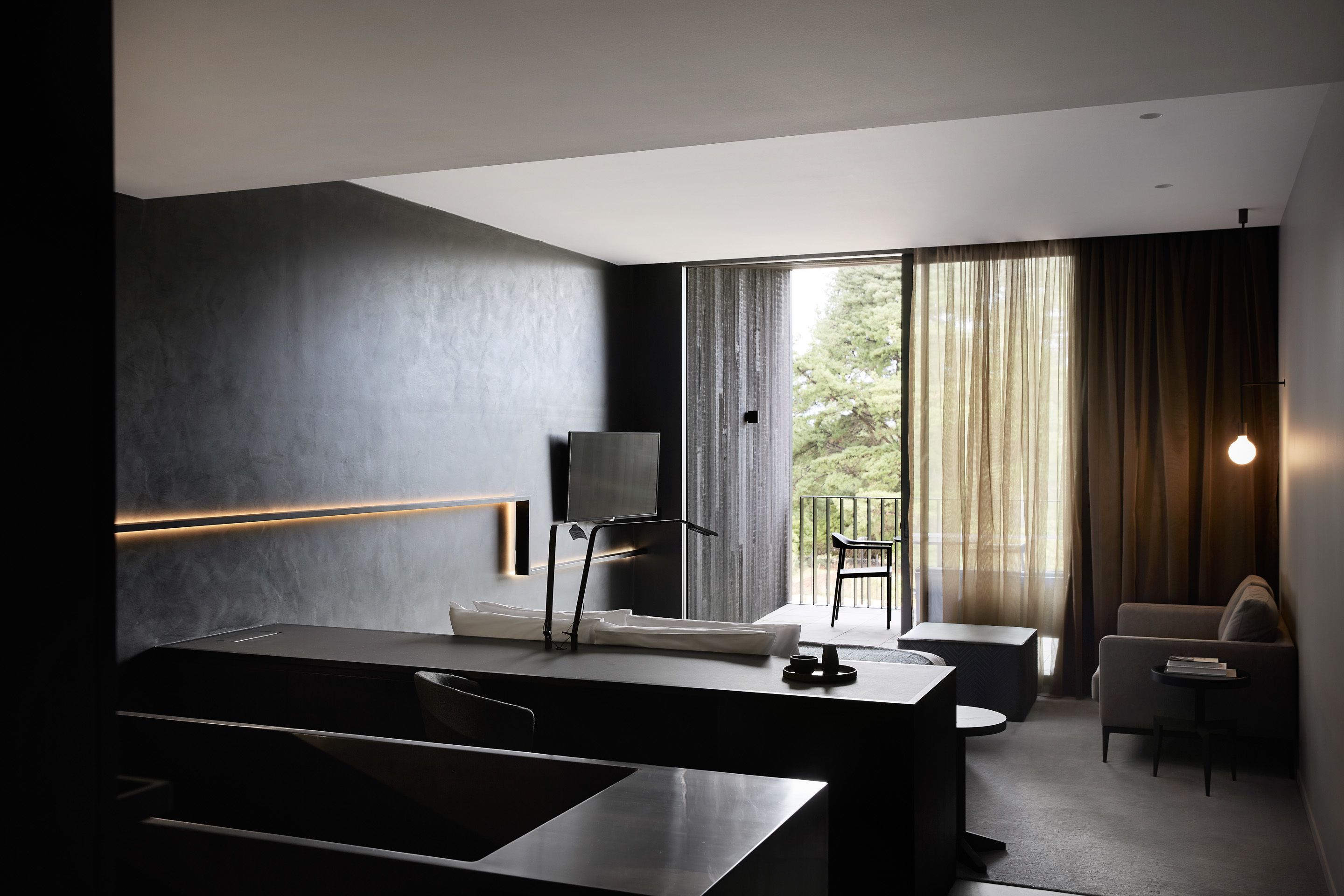 A modern hotel room with a large bed, a desk, and a TV. There is a balcony with a chair and curtains.