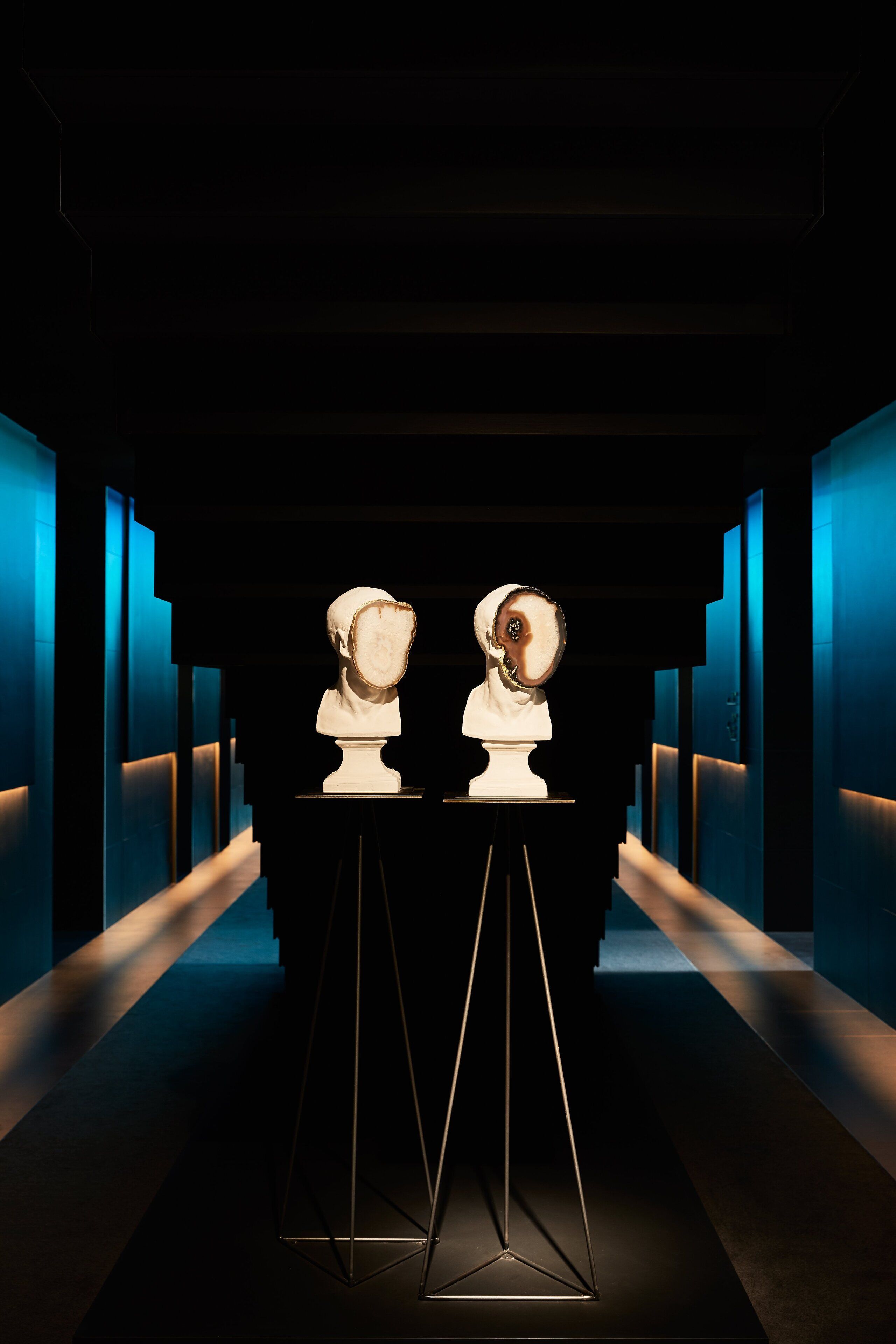 A hallway with two bust sculptures on pedestals, a dark floor, and blue illuminated walls.