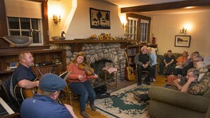 A cozy living room with a stone fireplace, wooden furniture, and a group of people engaged in a musical performance. - The Normaway Inn (Margaree Valley)