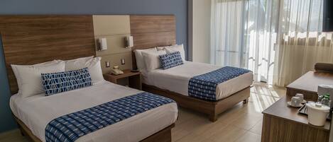 A hotel room with two beds, a wooden headboard, a bedside table with a lamp, a desk with a coffee maker, and a window with curtains.