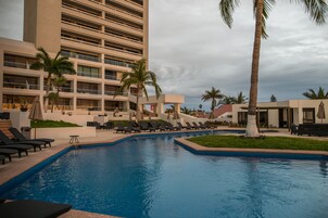 Piscina con sillas de descanso y palmeras, rodeada por un edificio de varios pisos con balcones.