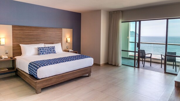 A modern hotel room with a large bed, bedside lamps, a nightstand, and a view of the ocean through floor-to-ceiling windows. - Ocean View Beach Hotel (Mazatlán)