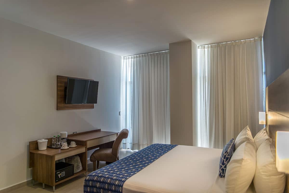 Una habitación de hotel con cama, escritorio, silla, televisión y cortinas.
