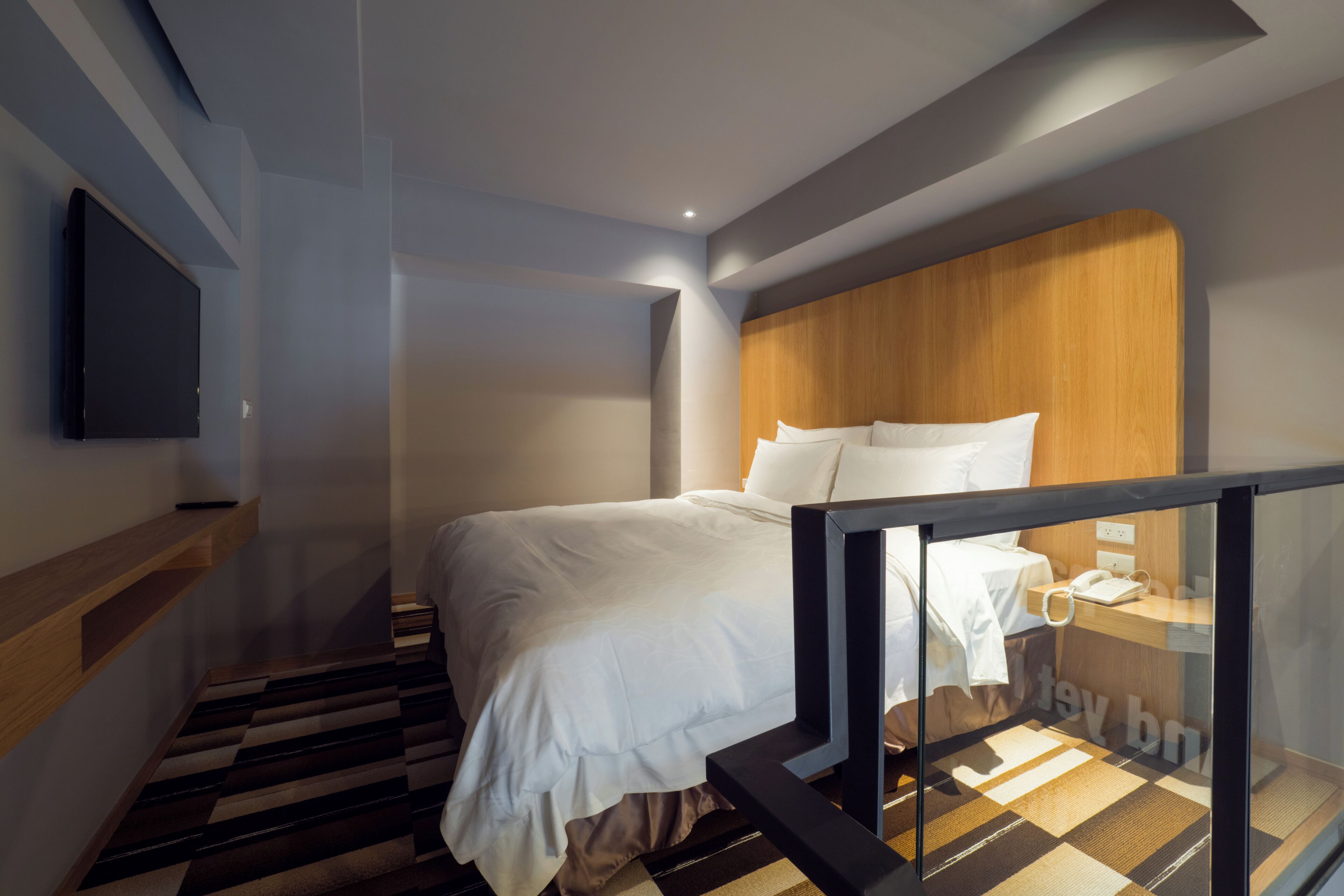 A modern hotel room with a large bed, a wooden headboard, a flat-screen TV mounted on the wall, and a staircase with a striped floor.