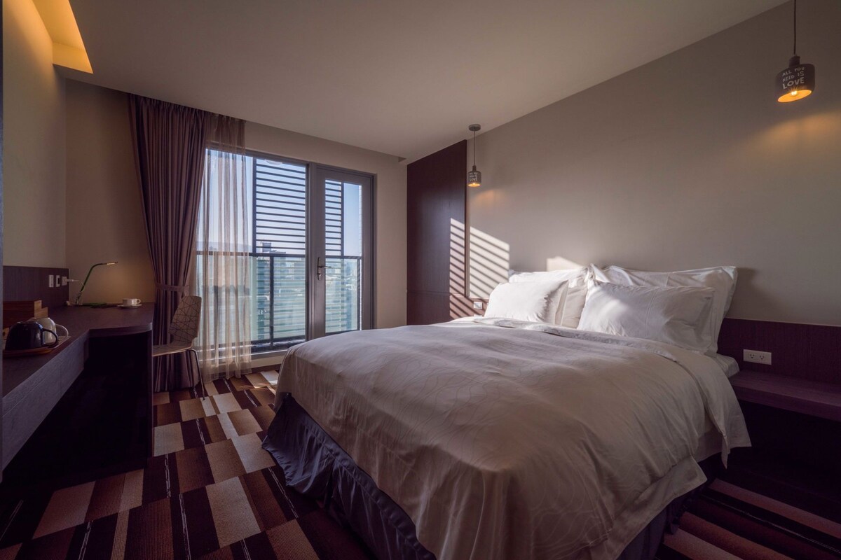 Una camera d'albergo con un letto grande, una scrivania con una lampada, una sedia e vista sulla città dalla finestra.